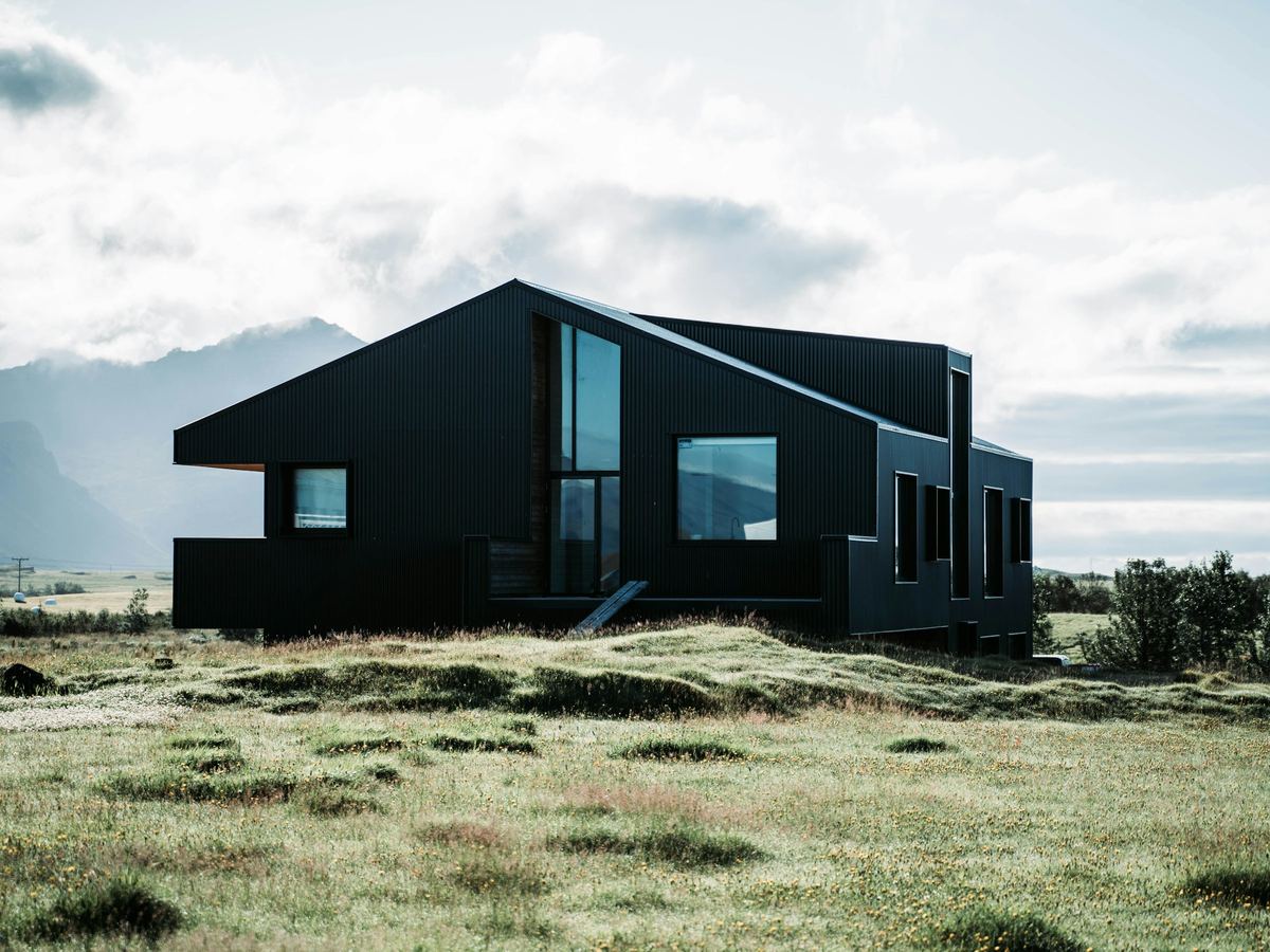 Dark timber-clad house with a pitched roof and large windows, set in open grassland below hills
